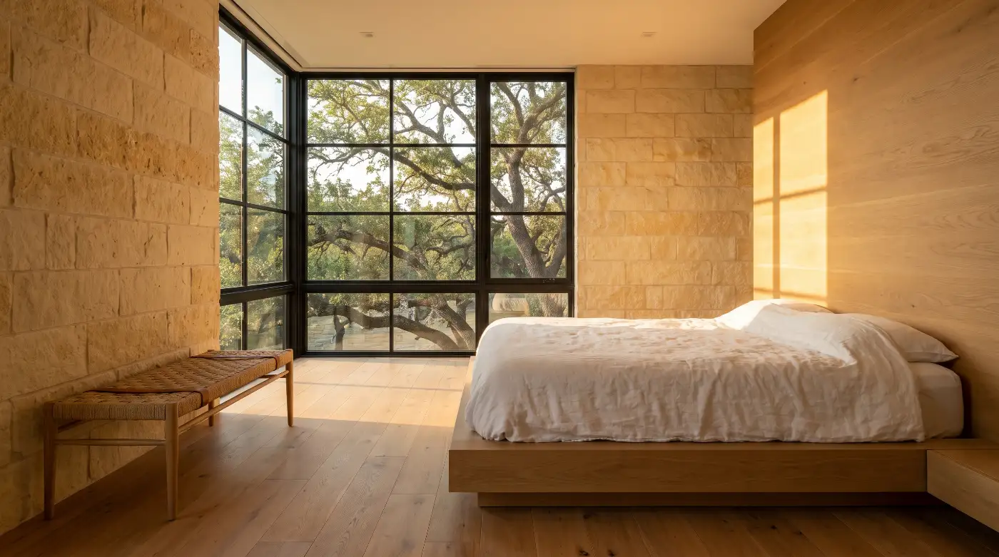 A primary bedroom with a limestone wall, a low white oak bed in white linen and a corner window filled with oak branches